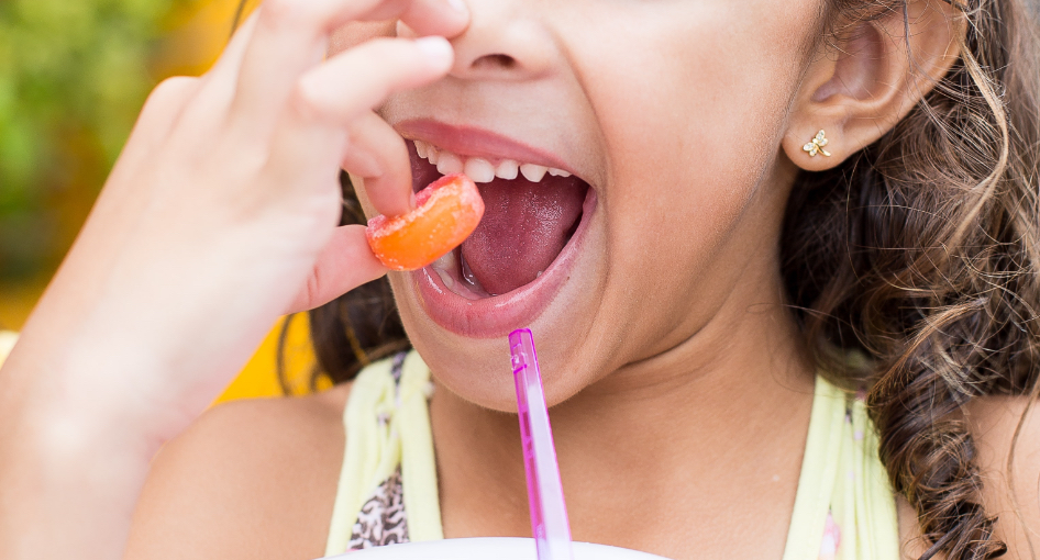 A person with curly hair holds an orange slice near their open mouth, with a purple straw in front.