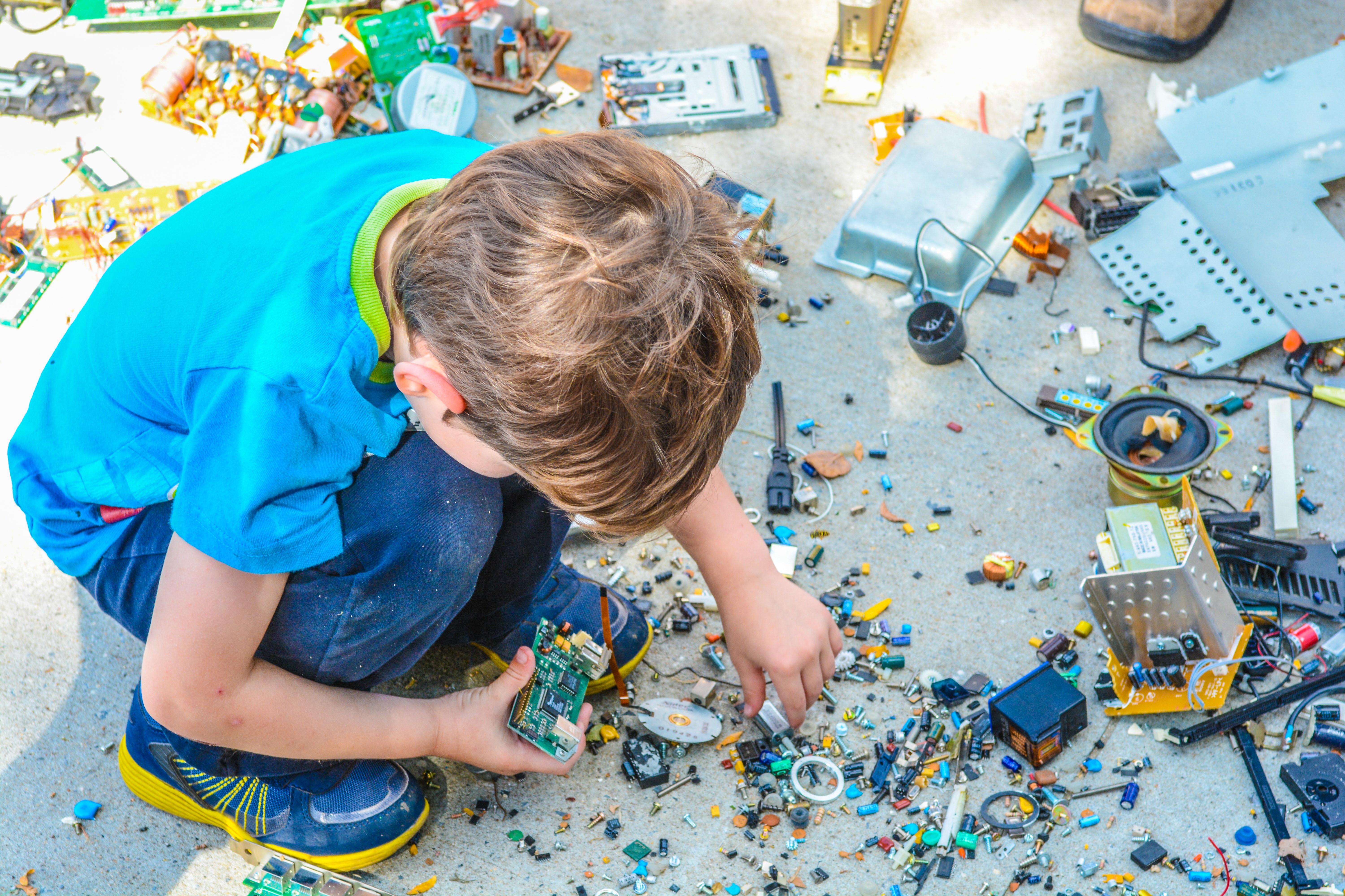 A child in a blue shirt crouches on concrete, holding a circuit board and pointing at small electronic components.