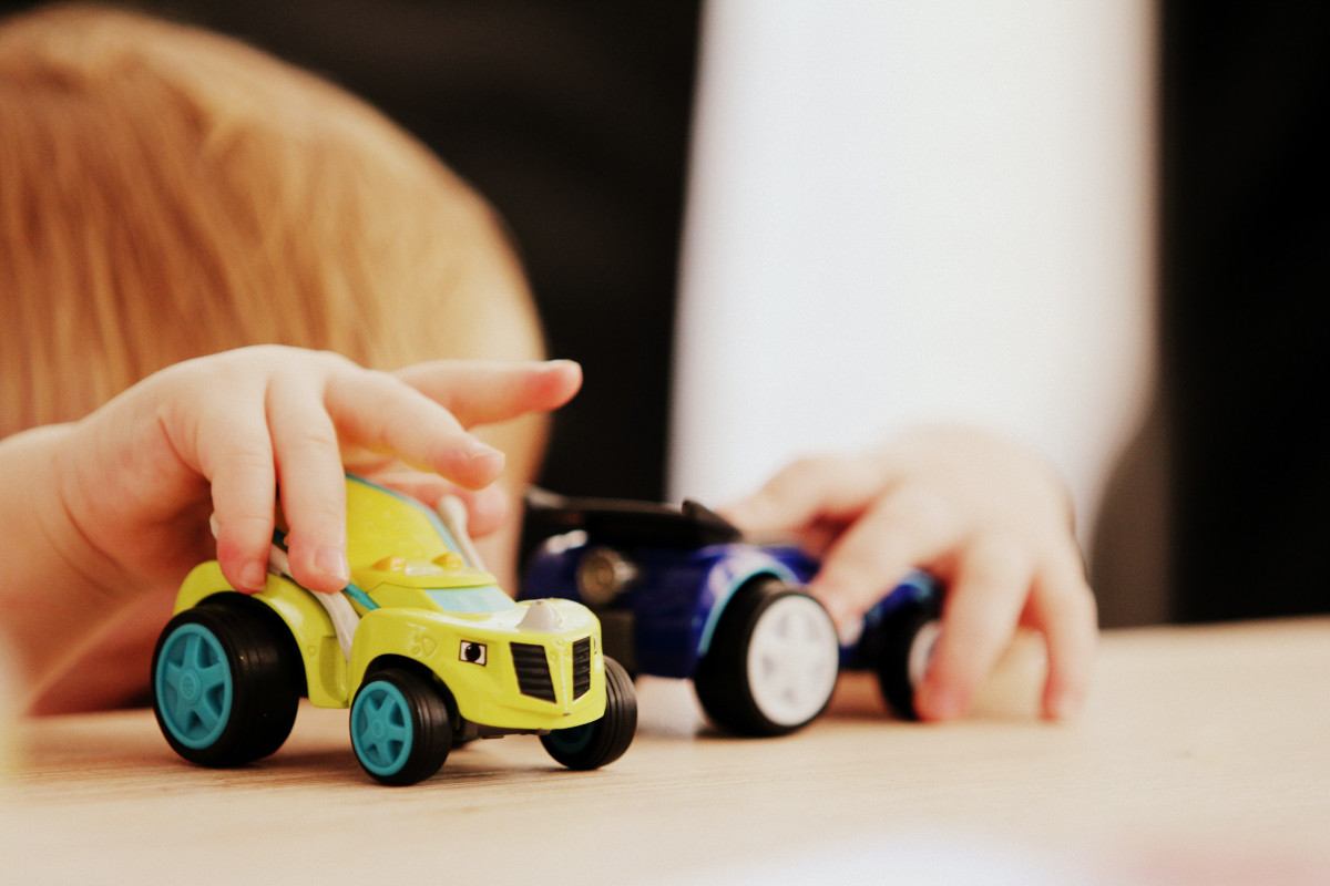A child's hands holding a yellow toy car with blue wheels and a blue toy car on a table.
