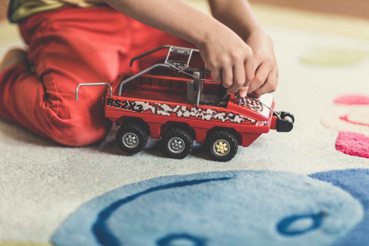 A child's hands adjust a red toy truck with 'RS2X3' and white patterns on a carpet.