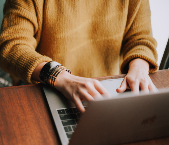 A person wearing a mustard yellow knit sweater types on a silver laptop placed on a wooden table, with multiple bracelets on their wrist.