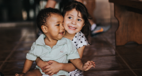 Two children sitting on a tiled floor, a girl in a white polka-dot shirt hugging a boy in a light green polo shirt.